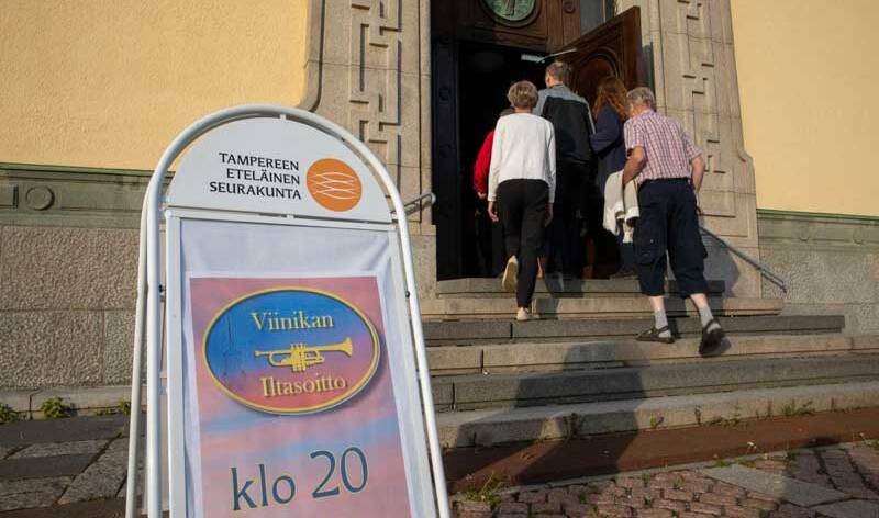 Ihmisiä menossa Viinikan kirkon ovesta sisään. Etualalla iso kyltti, jossa lukee Viinikan iltasoitto.