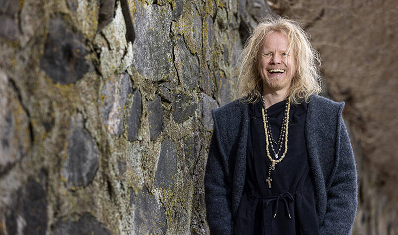 Virnistävä mies, jolla on pitkähkö liehuva tukka, kaulassaan koruja ja päällään naruvyöllä sidottu kaapu, seisoo vanhan kiviseinän vieressä.