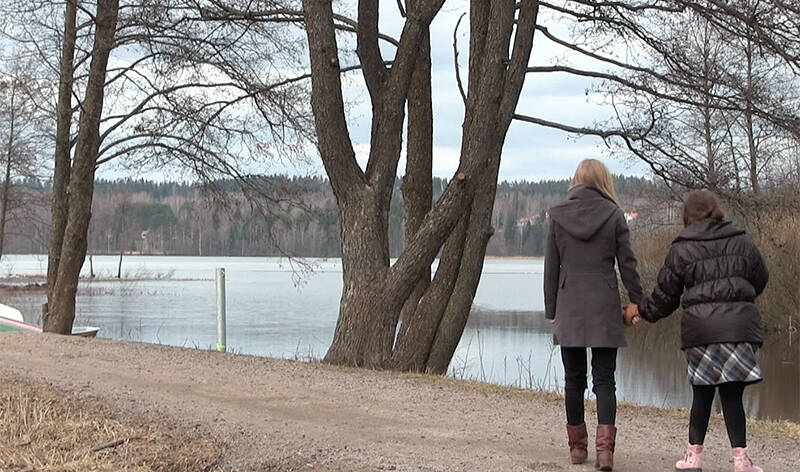 Syksyisellä hiekkatiellä järven rannan tuntumassa kävelee kaksi naista selin kameraan päin pitäen toisiaan kädestä kiinni.