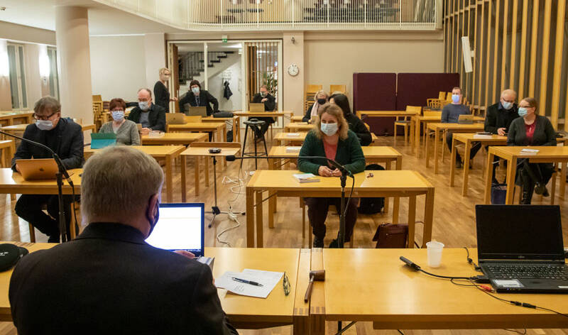 Salissa, joka näkyy etualalla olevan henkilön selän takaa, istuu noin kymmenen henkilöä, jokainen oman pöytänsä takana, ja jokaisella on kasvomaski.