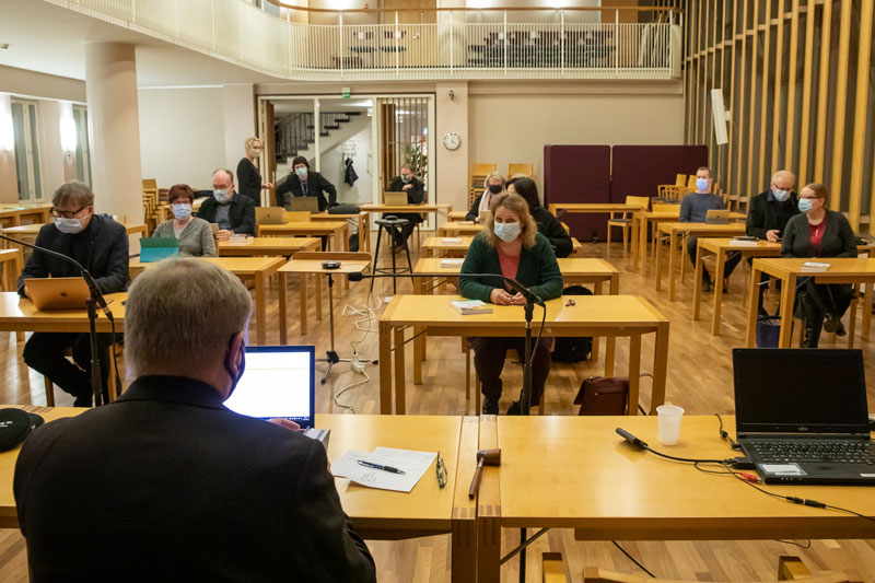 Salissa, joka näkyy etualalla olevan hemkilön selän takaa, istuu noin kymmenen henkilöä, jokainen oman pöytänsä takana, ja jokaislla on kasvomaski.