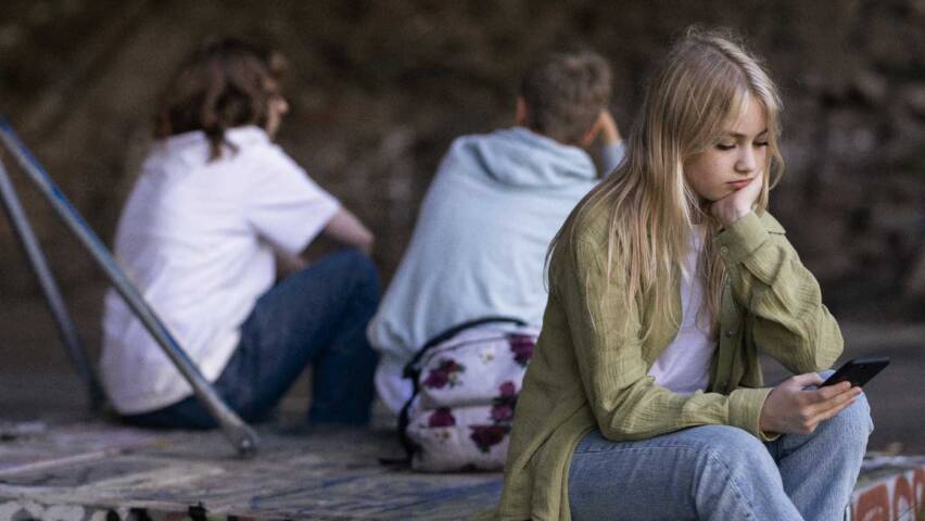 Nuori tyttö istuu lavan reunalla ja selaa kännykkää, hänen takanaan on pari ihmistä, jotka ovat kääntäneet hänelle selkänsä.