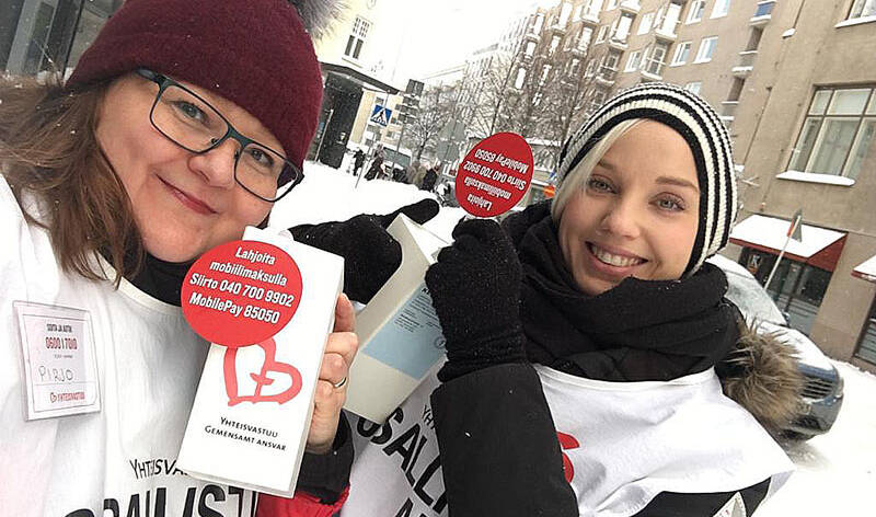 Kaksi naista hymyilee keräyslippaiden kanssa lumisessa katunäkymässä.