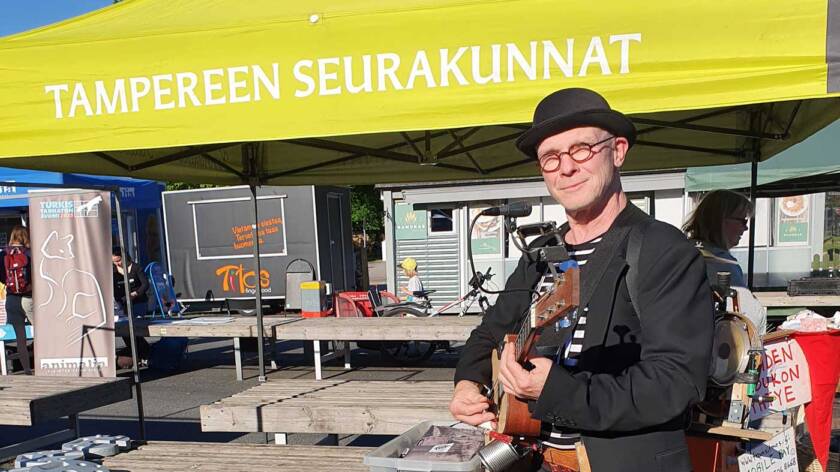 Knallipäinenmies ja kitara seisovat Tampereen seurakuntien torikatoksen edessä aurinkoisena kesäpäivänä.