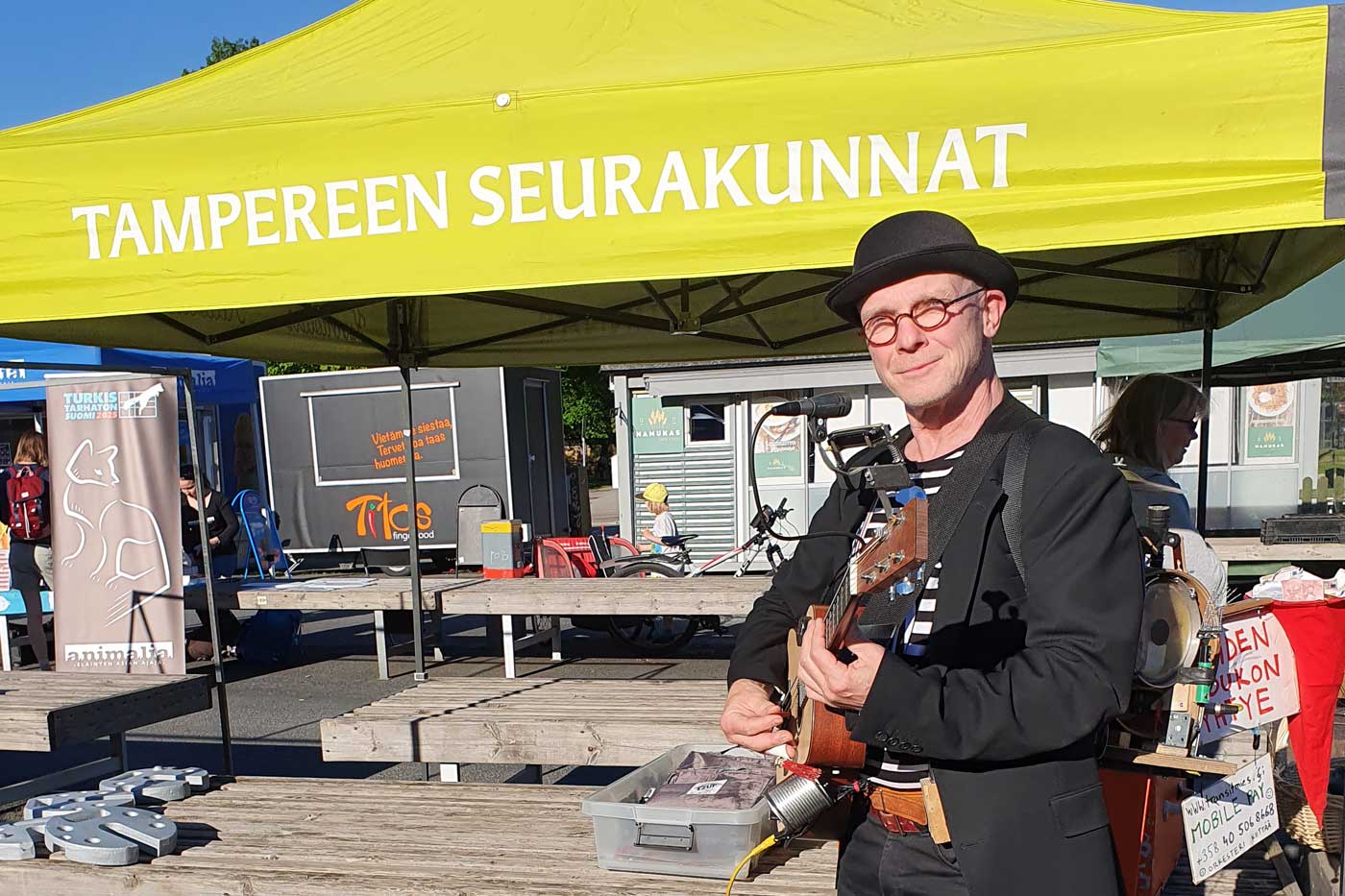 Knallipäinenmies ja kitara seisovat Tampereen seurakuntien torikatoksen edessä aurinkoisena kesäpäivänä.