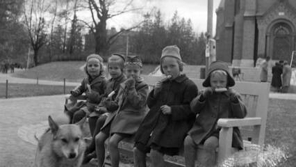Vanha kuva viidestä lapsesta, jotka istuvat penkillä syömässä jäätelöpuikkoja. Edessä on koira ja taustalla kirkko.