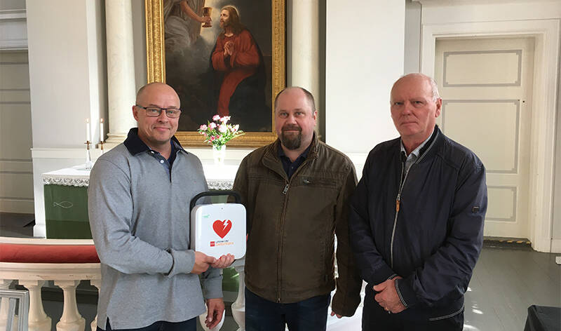 Kolme miestä seisoo kirkon alttarin edustalla. Kaksi miehistä on lahjoittanut kirkolle sydäniskurin, jonka on vastaanottanut kuvan kolmas mies.