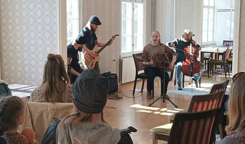 Bändi soittaa valoisassa tilassa. Etualalla on ihmisiä selin istumassa tuoleissa.