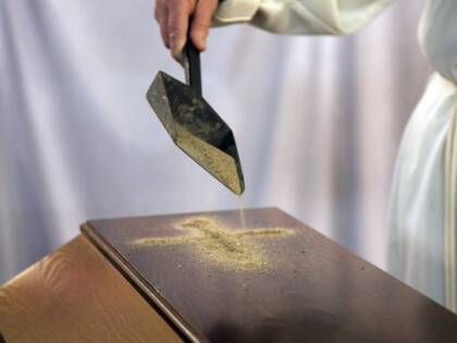 With a small shovel, sand is poured onto the top of the brown wooden coffin.