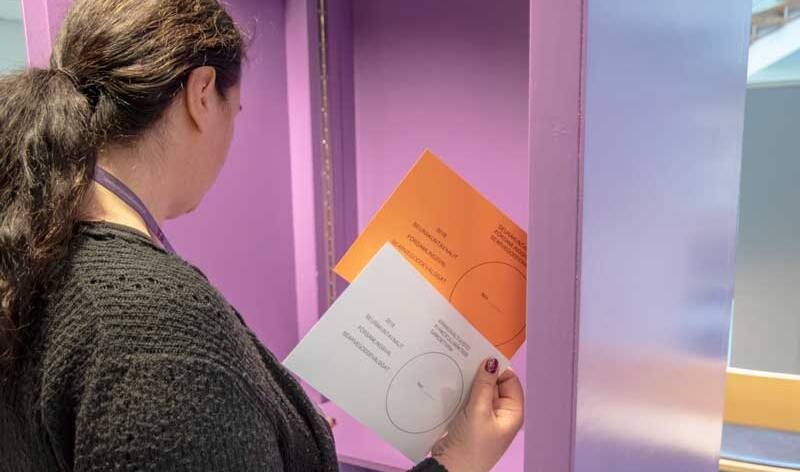 Pitkähiuksinen naisen seisoo äänestyskopissa ja pitää oikeassa kädessään orannsia ja valkoista äänestyslappua