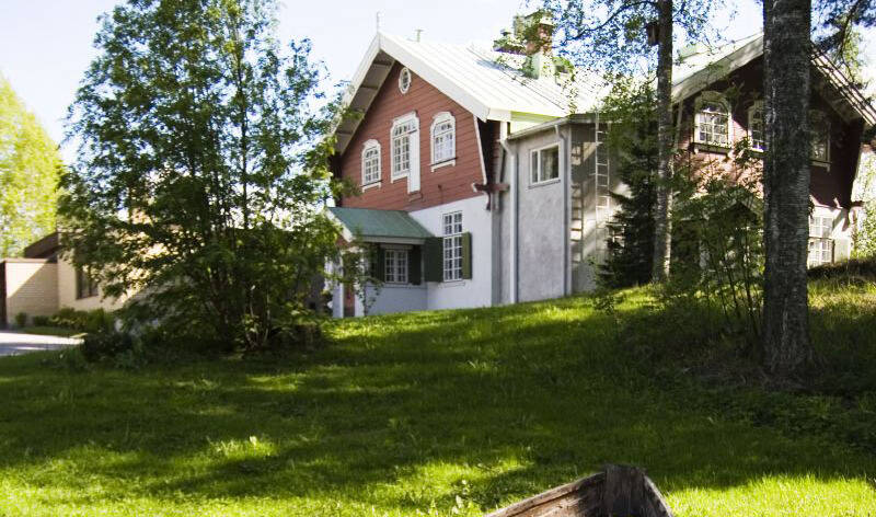 Mäen päällä on punavalkoinen puinen huvilarakennus, ja sen etualla on vihreä nurmialue,  jossa  kasvaa lehti- ja havupuita.