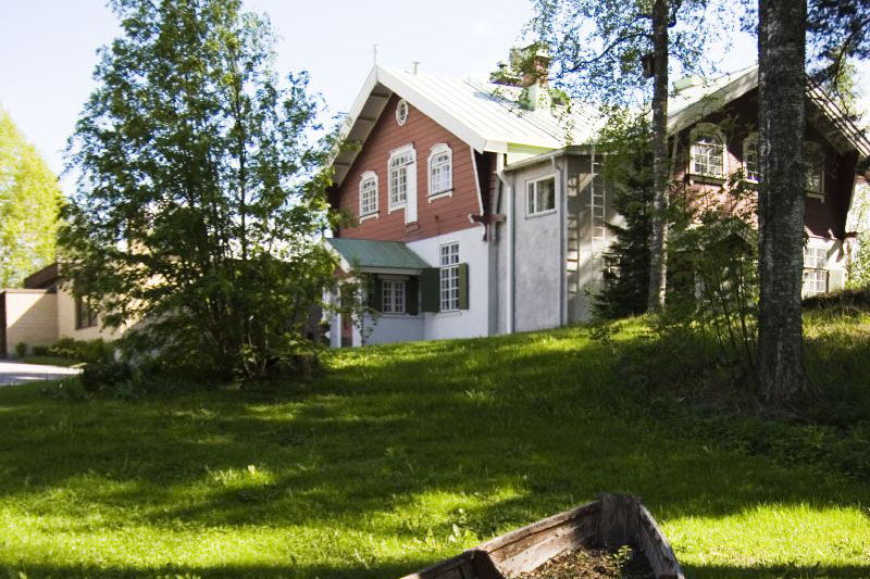 Mäen päällä on punavalkoinen puinen huvilarakennus, ja sen etualla on vihreä nurmialue,  jossa  kasvaa lehti- ja havupuita.