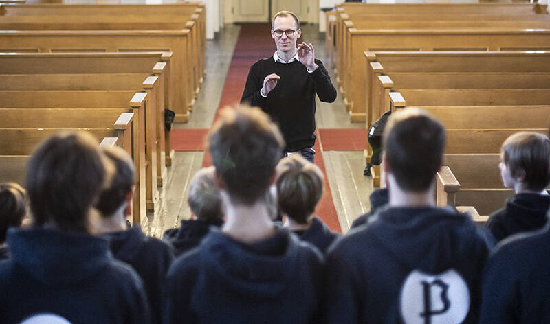 Nuorehko mies nostelee käsiään kirkon keskikäytävällä. Kuvassa etualalla on nuorukaisia selin katsojaan. Taustalla näkyvät urut.