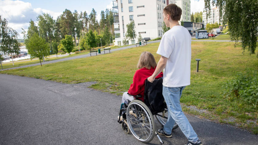 Nuori henkilä työntää pyörätuolia, jossa istuu harmaatukkainen vanhus, kesäinen järvimaisema on taustalla