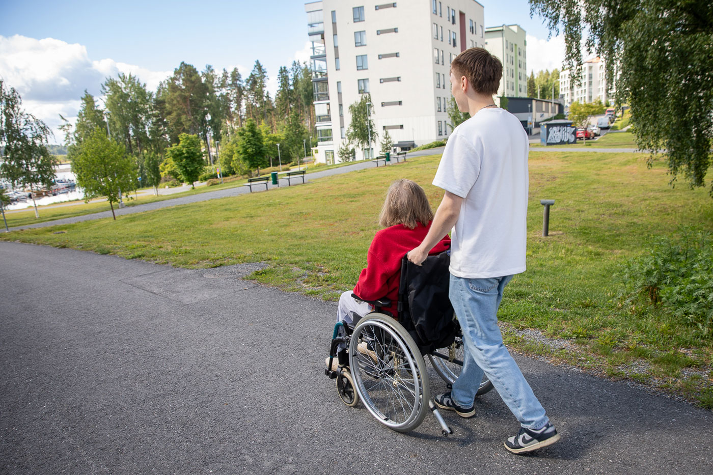 Nuori henkilä työntää pyörätuolia, jossa istuu harmaatukkainen vanhus, kesäinen järvimaisema on taustalla