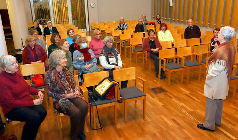 Nainen puhuu mikrofooniin ja hänen edessään on yleisöä, jotka istuvat tuoleilla.