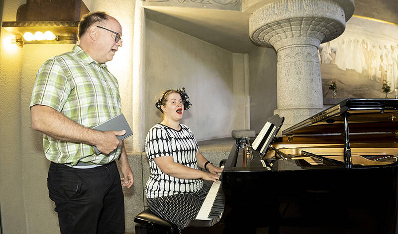 Mies laulaa seisoen ja nainen istuen pianon ääressä. Miehellä on oikeassa kädessään kiinni oleva kirja.