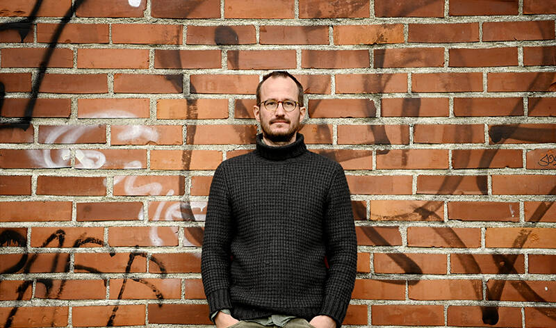 Elokuvaohjaaja Juha Kuosmanen seisoo kädet taskussa urbaani tiiliseinä taustallaan.