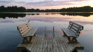 Auringon lasku heijastuu pilvistä järven pintaa violettin sävyin, ja järvessä on puinen laituri.
