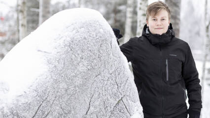 Nuorehko ihminen seisoo lumisessa maisemassa. Puolet kuvasta täyttää lumen peitossa oleva iso kivi.