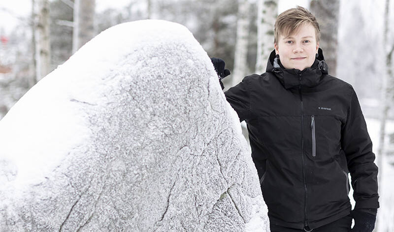 Nuorehko ihminen seisoo lumisessa maisemassa. Puolet kuvasta täyttää lumen peitossa oleva iso kivi.