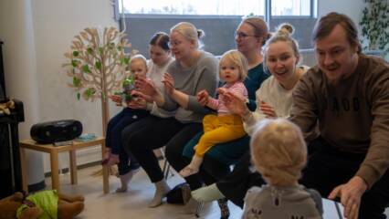 Ryhmä aikuisia ja lapsia istumassa sisätiloissa, osallistuen yhteiseen lauluhetkeen. Aikuiset ja lapset taputtavat käsiään.