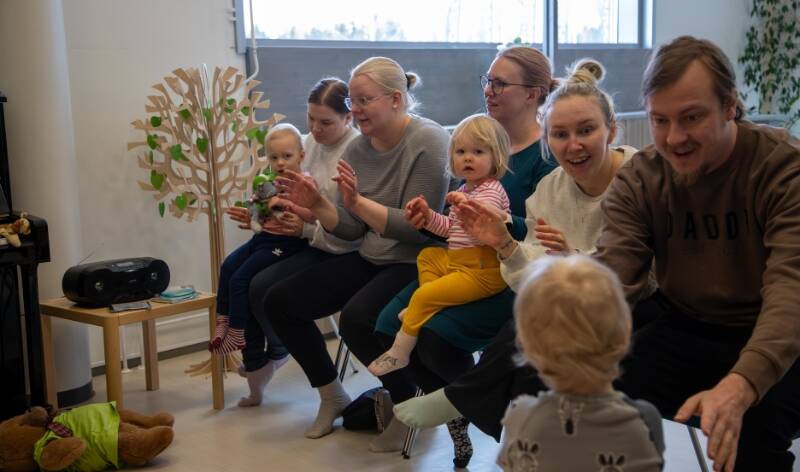 Ryhmä aikuisia ja lapsia istumassa sisätiloissa, osallistuen yhteiseen lauluhetkeen. Aikuiset ja lapset taputtavat käsiään.