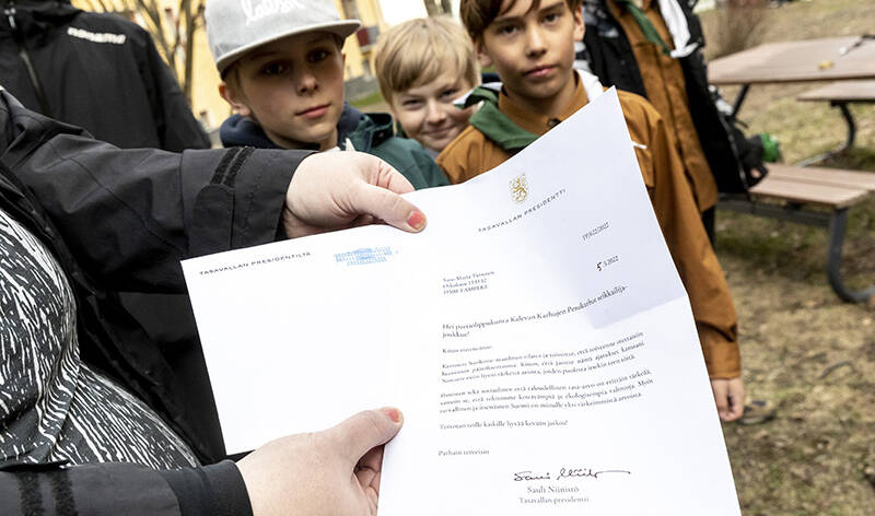 Jonkun käsissä on paperi, jossa on tekstiä. Takana näkyy kaksi pikkupoikaa.