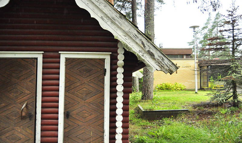 Etualalla osa puisesta aitasta, jossa on kaksi ovea. Taustalla tasakattoinen tiilirakennus, jonka seinässä lukee Ilkko.