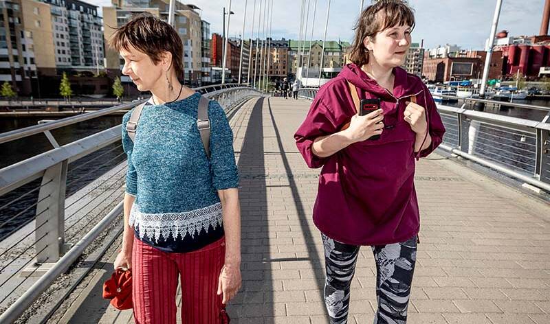 Kaksi naista kävelee sillalla joka ylitää vesistön. Taustalla taloja, tehdas ja laiturissa veneitä.