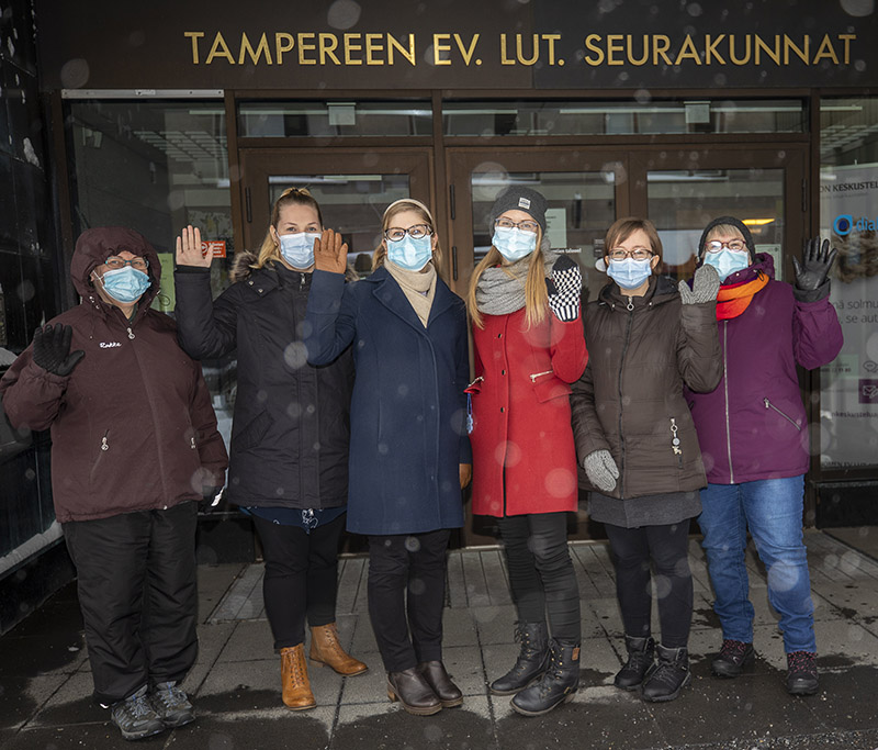Kuusi ihmistä seisoo maskit naamalla lumisateessa Seurakuntien talon edessä.