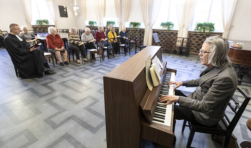 Etualalla mies soittaa kosketinsoitinta avarassa salissa, takana istuu ihmisiä tuoleilla.