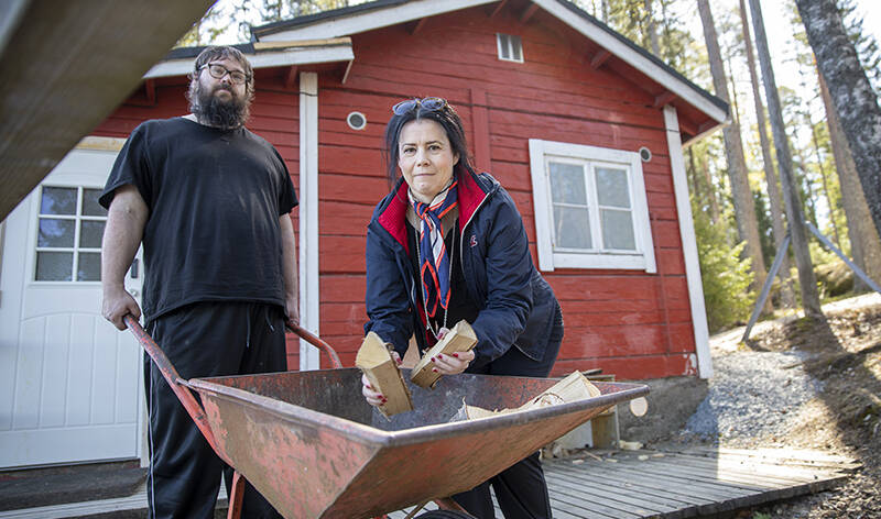 Yksi ihminen työntää kottikärryjä ja toinen laittaa siihen klapeja. Taustalla on tuparakennus.