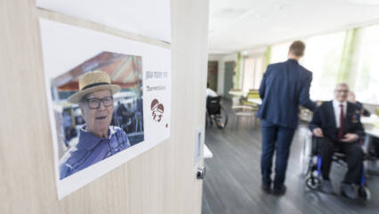 Puolet kuvasta täyttää vaalea ovi, johon on kiinnitetty paperinen tuloste. Tulosteessa on valokuva, jossa on vanhahko mies päässään olkihattu, ja paperissa on myös teksti Jalo-vaari 95v. Tervetuloa! Oven toisella puolen on sali, jossa nuorehko mies seisoo selin katsojaan ja kaksi muuta henkilöä istuu pyörätuolissa.