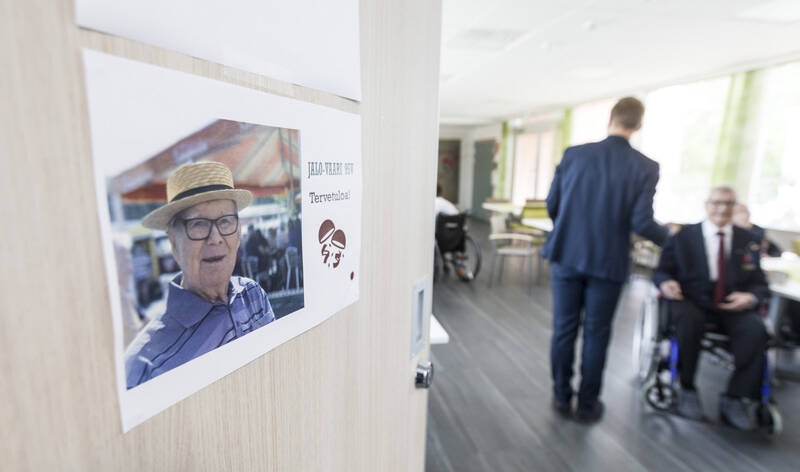 Puolet kuvasta täyttää vaalea ovi, johon on kiinnitetty paperinen tuloste. Tulosteessa on valokuva, jossa on vanhahko mies päässään olkihattu, ja paperissa on myös teksti Jalo-vaari 95v. Tervetuloa! Oven toisella puolen on sali, jossa nuorehko mies seisoo selin katsojaan ja kaksi muuta henkilöä istuu pyörätuolissa.
