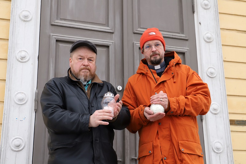 Kaksi miestä seisoo kirkon edustalla sytytetyt kynttilät käsissään.