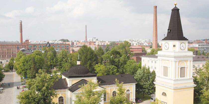 A summery aerial view of the light yellow wooden Old Church from the direction of the Central Square, with the bell tower to the right of the church, in the background green deciduous trees, and the red brick factory buildings of the Finlayson and Tampella areas with their chimneys.