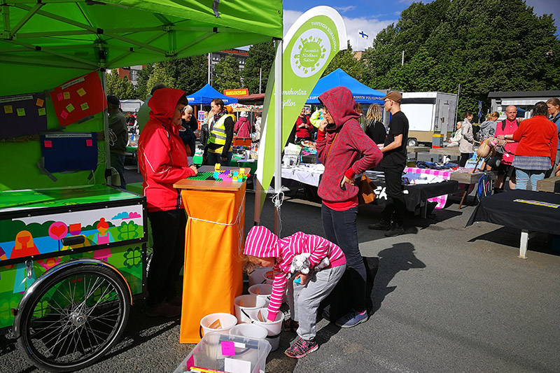 Katoksen edessä seisoo ihminen, jolle toinen ihminen juttelee. Katosta vasten on polkupyörän rengas. Lapsi tavoittelee jotain edessä olevista astioista. Taustalla on pöytiä, ihmisiä, pöytiä ja koppeja.