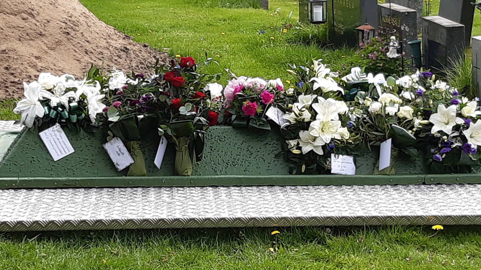 Hautakumpu, jonka päällä kukkalaitteita. Taustalla hiekkakasa ja nurmikkoa.