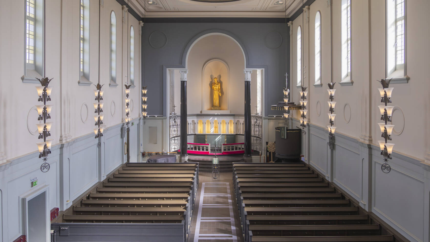 Fi: Kaunis kirkko, jossa on tyylikkäitä ikkunoita ja puupenkit. Alttari on keskiössä, ja seinillä on koristeellisia valaisimia. / Sv: En vacker kyrka med stiliga fönster och bänkar i trä. Altaret är i centrum, och väggarna har dekorativa lampor.