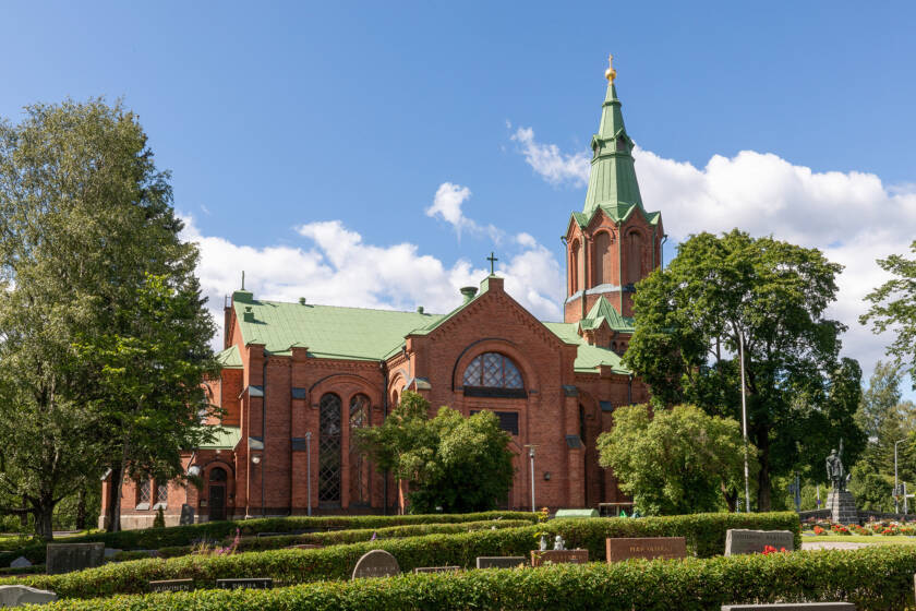 Punatiilinen kirkko vihreällä katolla, ympärillä puita ja pensaita, taustalla kirkas sininen taivas ja valkoisia pilviä.
