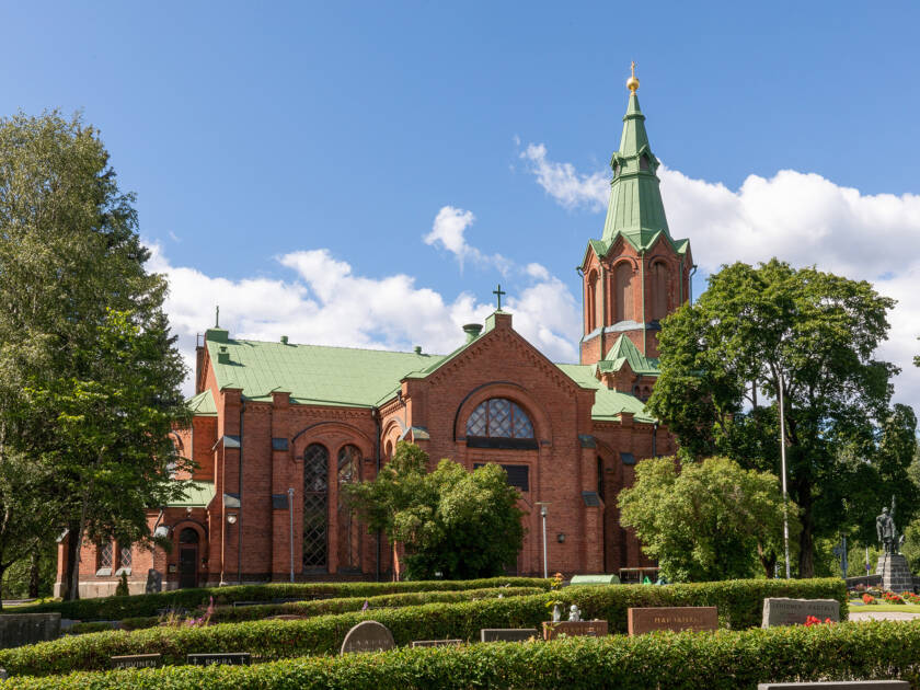 Punatiilinen kirkko vihreällä katolla, ympärillä puita ja pensaita, taustalla kirkas sininen taivas ja valkoisia pilviä.