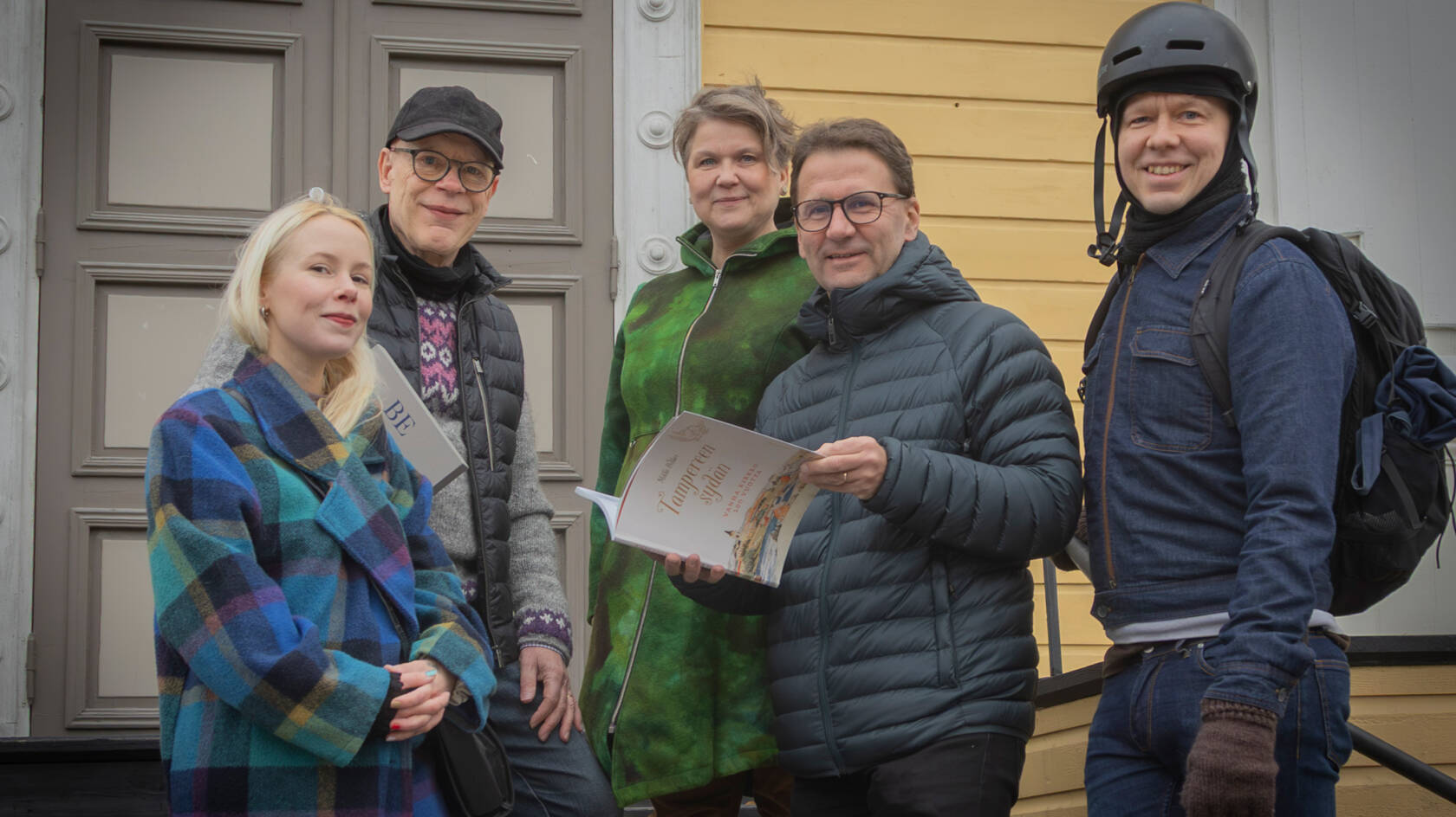 Viisi ihmistä seisoo ulkona keltaisen puukirkon portailla. Yhdellä on kädessään kirja ja kaikki hymyilevät.