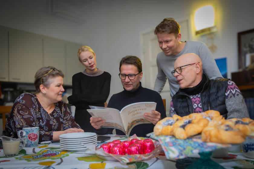 Viisi ihmistä tarkastelee avoinna olevaa kirjaa pöydän ääressä. Oikeanpuoleinen heistä osoittaa kädellään jotain kohtaa kirjasta. Pöydällä on herkkuja.