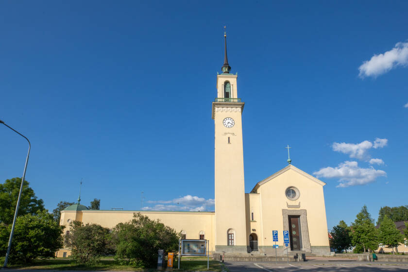 Kirkko, jossa on korkea torni ja kello, taustalla kirkas sininen taivas ja valkoisia pilviä. Alhaalla näkyy puistoalue.