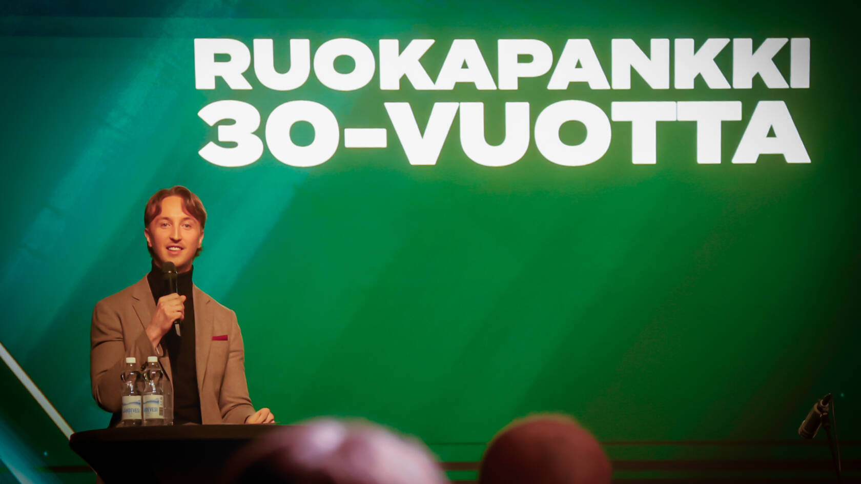 Ilmari Nurminen seisoo lavalla mikrofoni kädessä, taustalla teksti Ruokapankki 30-vuotta.