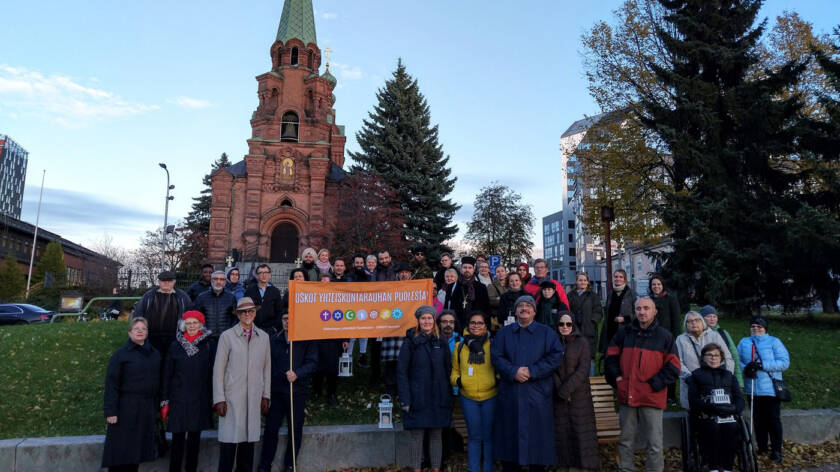Vuoden 2024 rauhankävelyn osallistujat seisovat Sorin aukiolla Tampereen ortodoksisen kirkon edessä. Ryhmän keskellä pidetään oranssia kylttiä, jossa lukee ‘Uskot yhteiskuntarauhan puolesta!’.