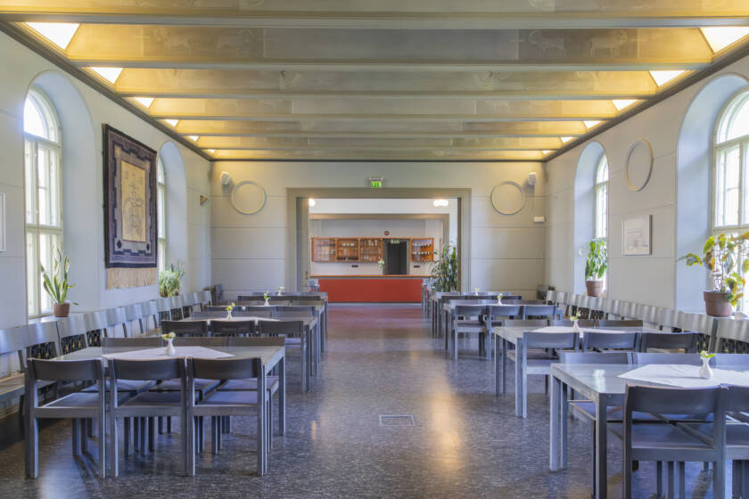 Fi: Tyhjät ruokasalit, joissa on harmaat pöydät ja tuolit. Ikkunoista tulvii luonnonvaloa, ja taustalla näkyy keittiö. / Sv: Tomma matsalar med grå bord och stolar. Naturligt ljus strömmar in genom fönstren, och köket syns i bakgrunden.