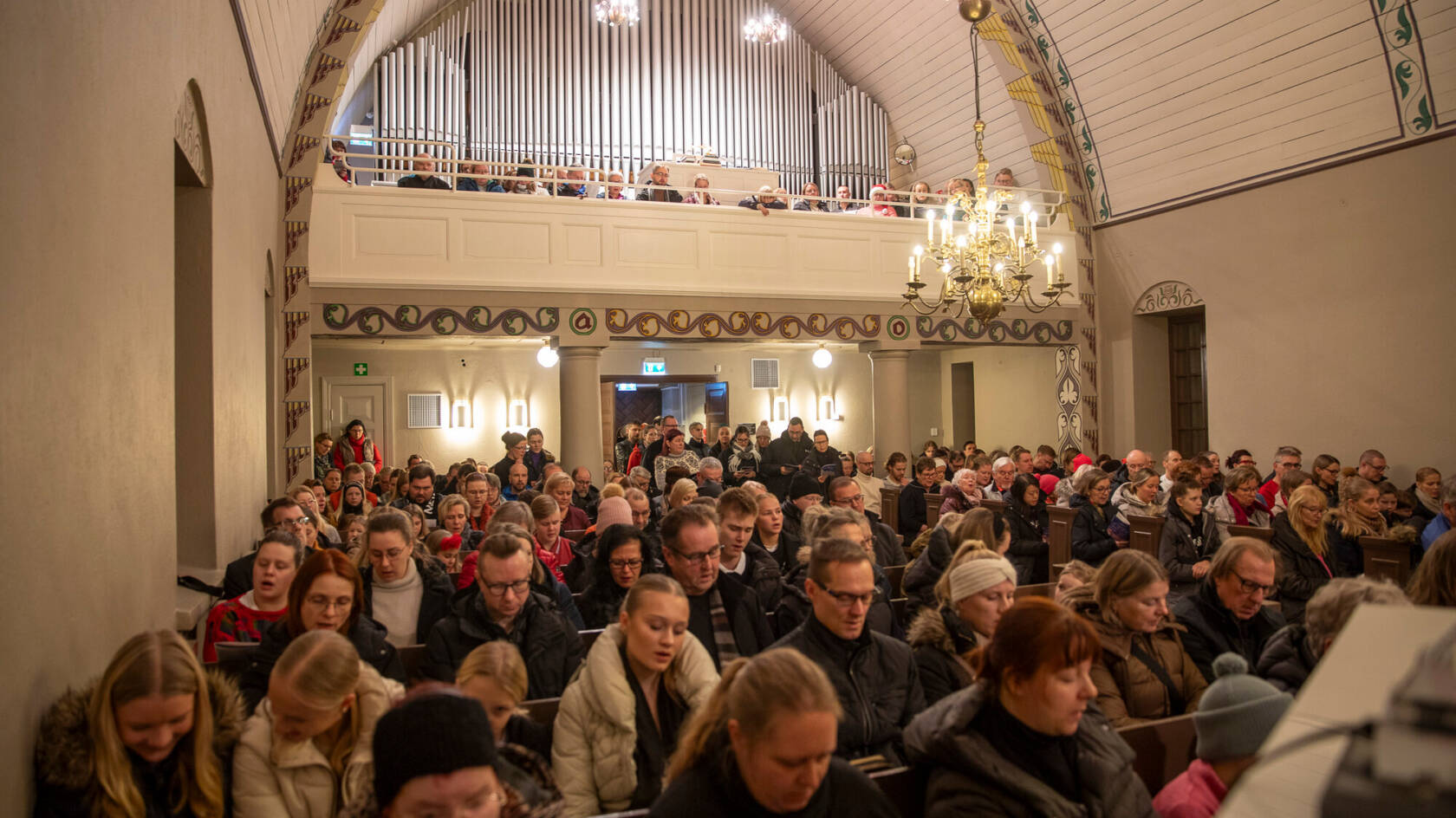 Ihmiset istuvat kirkossa, katsoen eteenpäin. Taustalla näkyy urut ja koristeelliset yksityiskohdat.