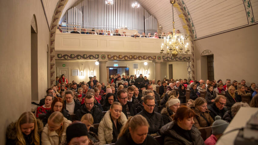 Ihmiset istuvat kirkossa, katsoen eteenpäin. Taustalla näkyy urut ja koristeelliset yksityiskohdat.